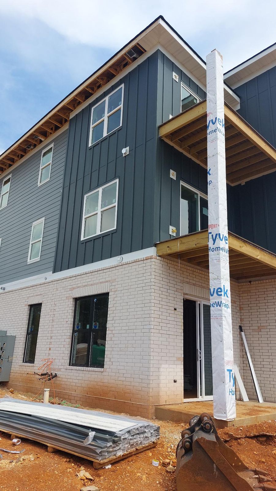 Multi-family building exterior with dark green board-and-batten siding over a brick base, balconies, and Tyvek-wrapped support post.