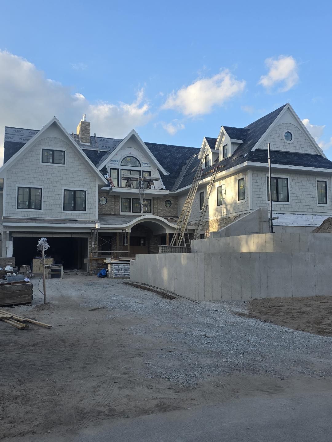 Large custom home with completed shake-style siding, stone accents, multiple gables, and a poured concrete retaining wall.