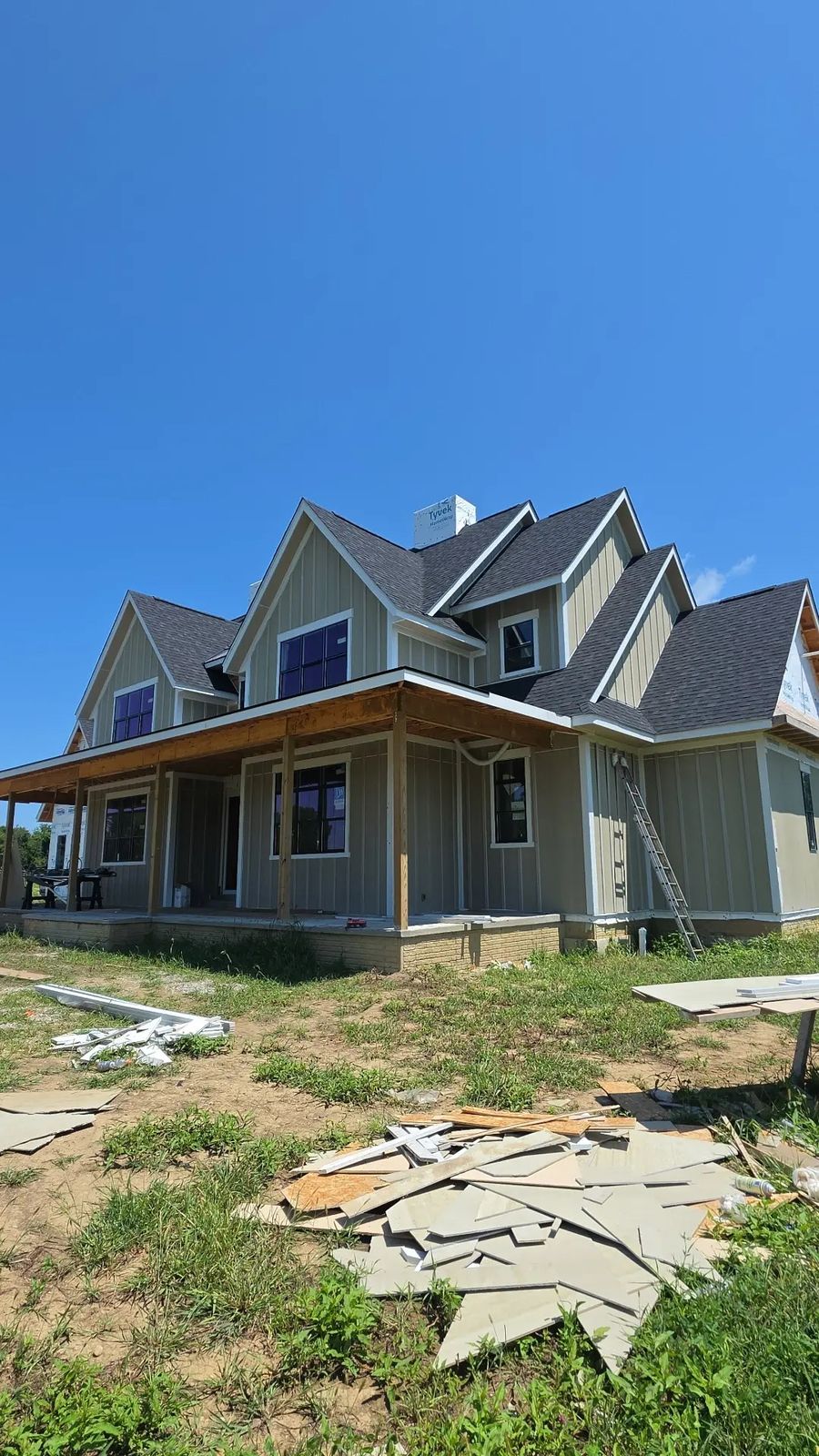 New construction farmhouse-style home with sage green board-and-batten siding and a wraparound covered porch, under a clear blue sky.